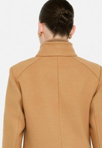 Cappotto color cammello con colletto a punta, texture liscia e vestibilità sagomata con cuciture delle spalle a vista.