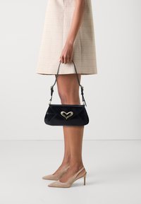 Svart lackläderhandväska med en guld hjärtformad detalj, smal axelrem och en strukturerad design. Bärs tillsammans med en ljus rutig klänning och bleka klackar.