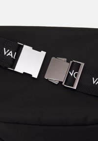 Schwarze Stofftasche mit strukturierter Oberfläche, ausgestattet mit metallischen Schnallen und verstellbaren Riemen mit einem Logo. Minimalistisches Design, kompakte Form.