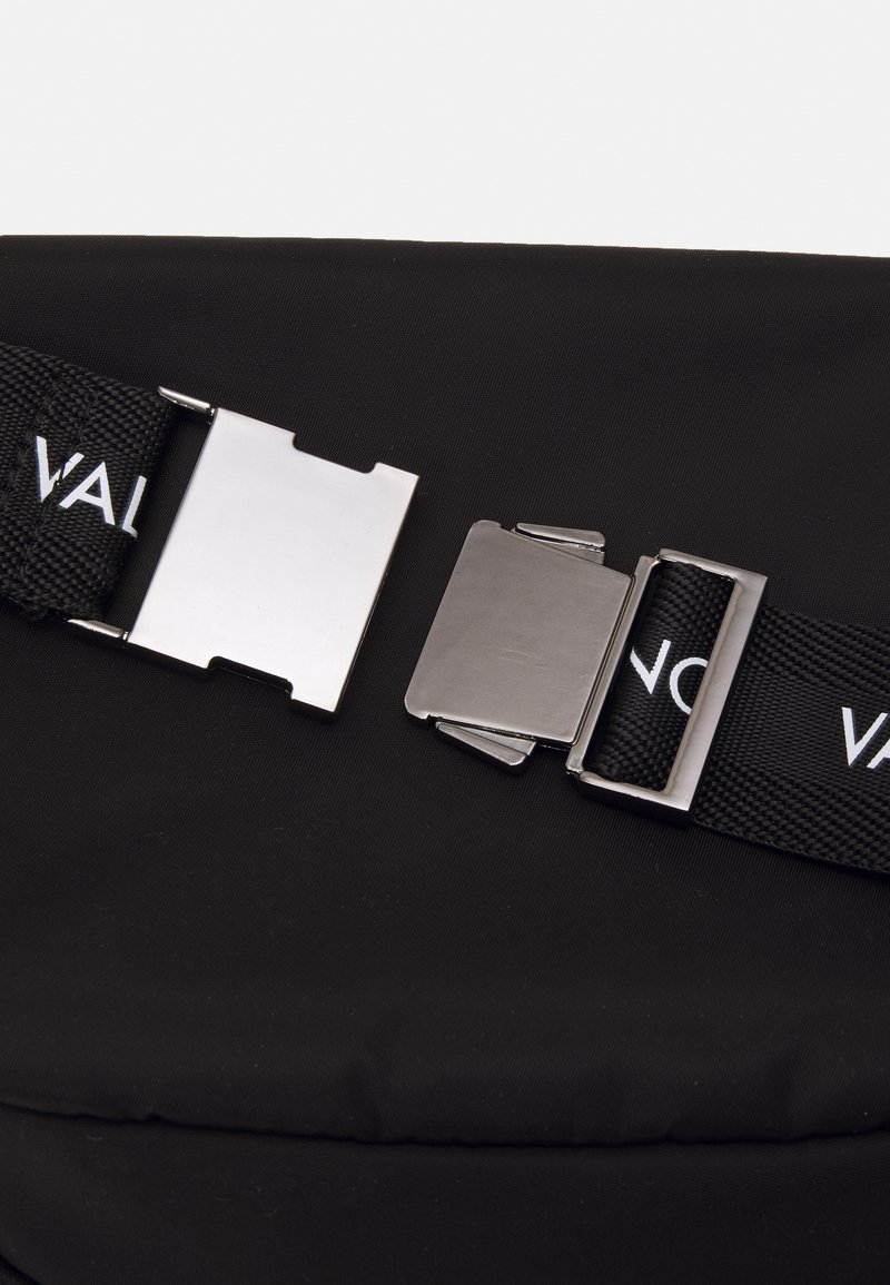 Sac en tissu noir avec une surface texturée, comportant des boucles métalliques et des sangles ajustables avec un logo. Design minimaliste, forme compacte.