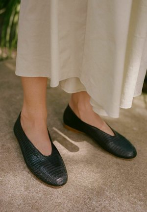 Scarpe nere basse con un motivo testurizzato, punta arrotondata e suola bassa marrone, abbinate a una gonna beige fluida.