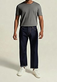 Homme portant un t-shirt gris à manches courtes, un jean bleu foncé retroussé et des baskets blanches, debout devant un fond uni.