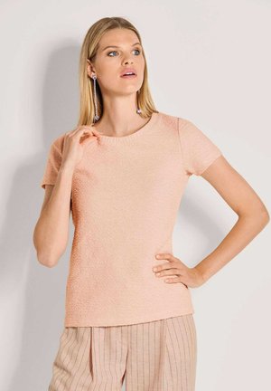 Pfirsichfarbenes Kurzarmhemd mit rundem Ausschnitt, kombiniert mit hell gestreifter Hose. Der Stoff hat ein dezentes Muster und wirkt weich.