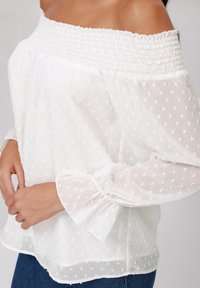 White off-the-shoulder blouse with smocked neckline and flared sleeves, featuring textured polka dot patterns and a lightweight, sheer fabric.