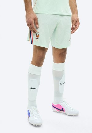 Joueur de football masculin portant un short vert clair de l'équipe de France avec l'emblème du coq, des chaussettes blanches Nike avec protège-tibias, et des crampons blancs avec des accents roses et bleus.