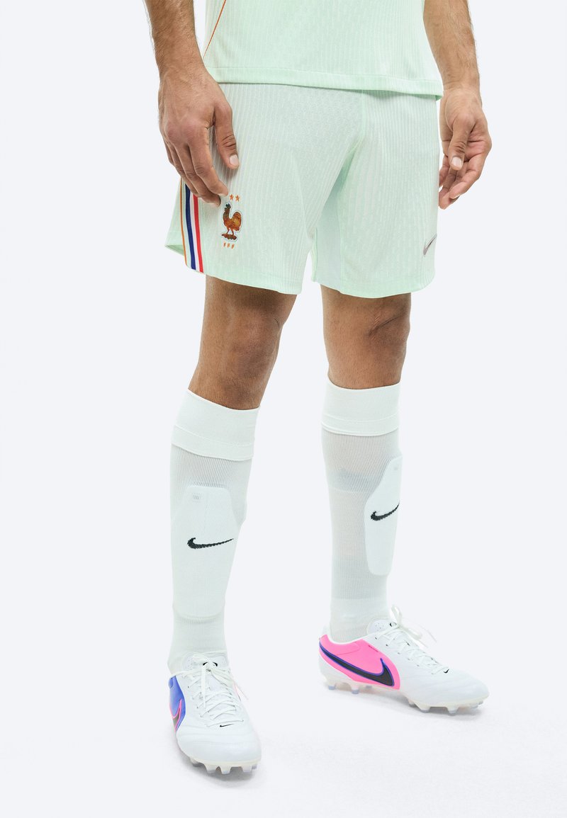 Calciatore maschio che indossa pantaloncini verde chiaro della Francia con emblema del gallo, calzini bianchi Nike con parastinchi e scarpe da calcio bianche con dettagli rosa e blu.