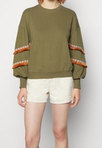 Felpa verde oliva con maniche oversized, caratterizzata da frange arancioni e una rifinitura floreale sulle maniche. Abbinata a pantaloni corti beige chiaro.