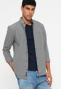 Schwarz-weiße karierte Hemd mit Kragen, getragen über einem navyblauen T-Shirt, kombiniert mit hellblauen Jeans.