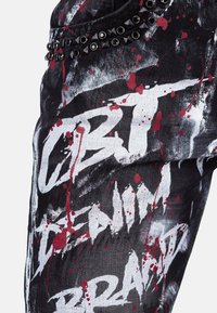 Pantaloni in denim nero con design a spruzzi di vernice bianca e rossa, hardware con borchie lungo la vita e elementi testuali grafici in evidenza.