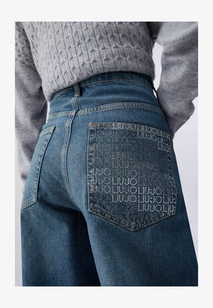 Krupan plan plavih traperica s džepom na stražnjoj strani ukrašenim cirkonima na kojima je ponovljen tekst "LIU JO", nošenih s sivim pletenim džemperom.