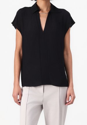 Woman wearing a black cap-sleeve blouse with a deep V-neck collar and light beige tailored pants, standing with arms relaxed by sides.