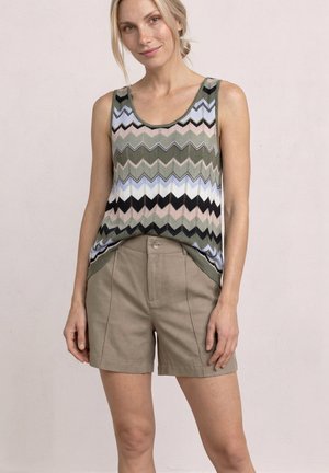 Kvinde iført en ærmeløs top med zigzag-mønster i grøn, sort og hvid med beige højtaljede shorts, stående mod en ensfarvet baggrund.
