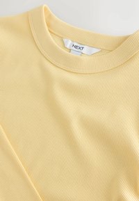 Hellgelbes, geripptes T-Shirt mit rundem Ausschnitt und einer subtilen Textur, versehen mit einem kleinen Etikett mit dem Markennamen "NEXT."