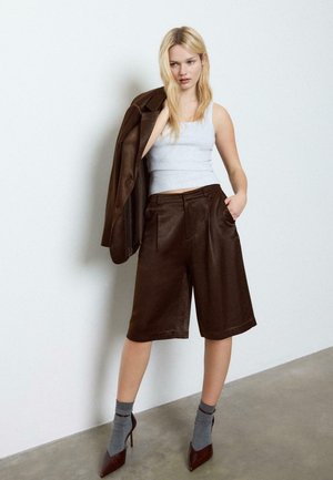 Femme blonde portant un short ample marron, une veste assortie jetée sur l'épaule, un haut blanc sans manches, des chaussettes grises et des talons hauts marron.