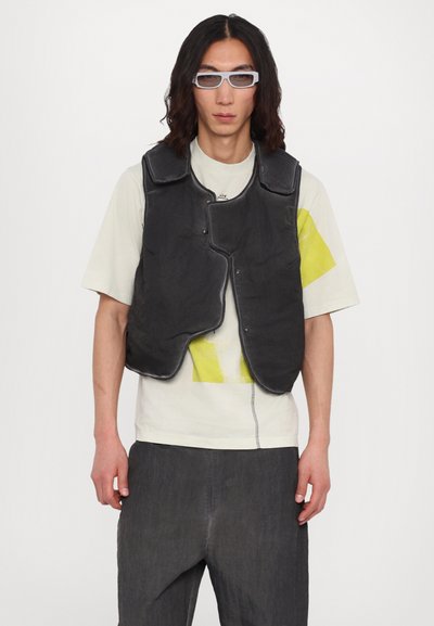 Gilet noir matelassé aux bords arrondis et fermetures à pression ; porté sur un T-shirt gris clair avec un design jaune ; matériau texturé et léger.