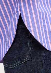 Μπλε και ροζ ριγέ άκρη πουκάμισου κρέμεται πάνω από την τσέπη ενός σκούρου μπλε τζιν με ορατά λευκά ραφτικά.