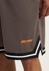 Szorty sportowe z szarego materiału o siateczkowej fakturze. Wykończone czarno-białym paskiem, z pomarańczowym logo "URTRD SPORT" na brzegu.