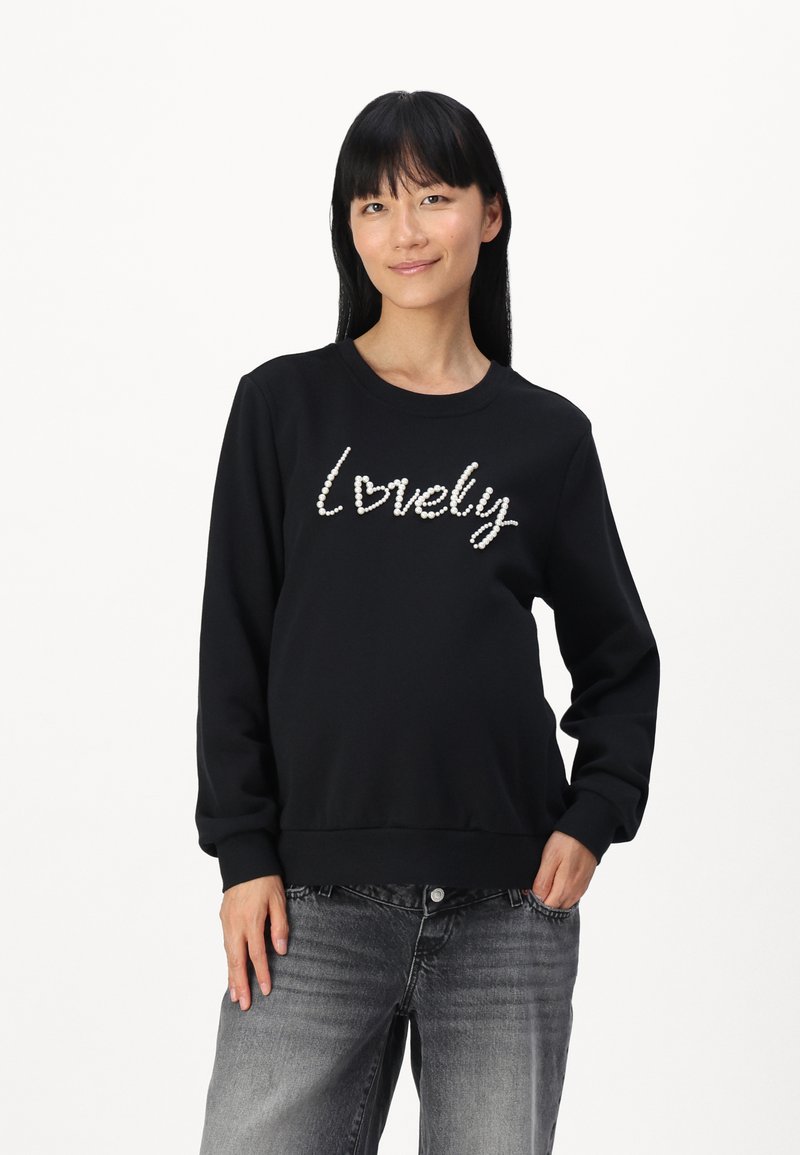 Sweatshirt noir avec le texte "lovely" orné de sequins blancs. Avec un col rond et des poignets côtelés. Porté avec un jean gris.