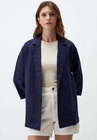Blazer oversize in lino navy con colletto sciallato, apertura frontale e maniche a tre quarti, abbinato a una maglietta beige e pantaloni corti bianchi.