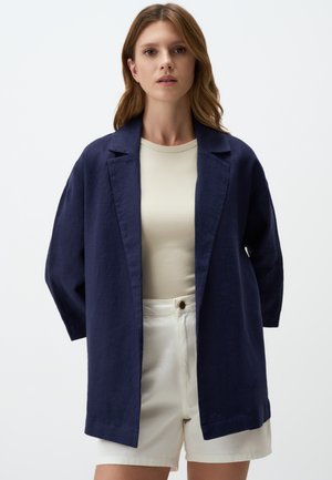 Marineblaues Leinen-Übergröße-Blazer mit Schalkragen, offener Front und Dreiviertelärmeln, kombiniert mit einem beigen Oberteil und weißen Shorts.