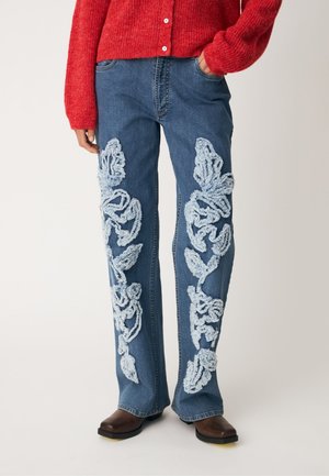 Jean en denim avec de grands motifs floraux texturés bleu clair sur les jambes avant, porté avec des chaussures marron et un pull en tricot rouge.