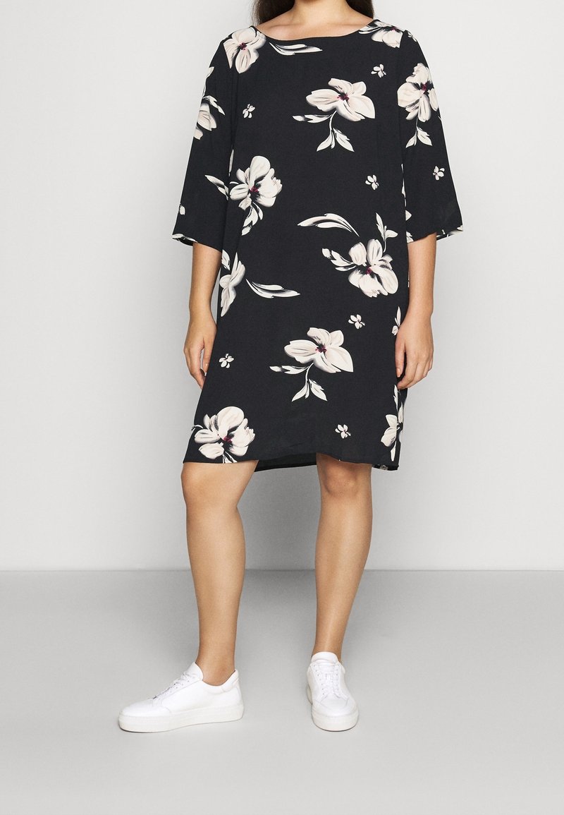 Femme portant une robe noire au genou avec motif floral blanc et manches au coude, associée à des baskets blanches, debout devant un fond uni.