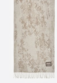 Beige geweven deken met een gestructureerd patroon in lichtere en donkere tinten, afgewerkt met franjes. Beschikt over een merklabel in de hoek.