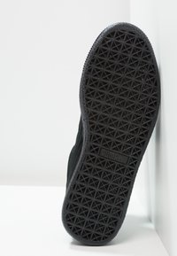 Semelle de chaussure de sport noire avec motif de bande de roulement géométrique et logo Puma en relief au centre, inclinée sur un fond blanc.