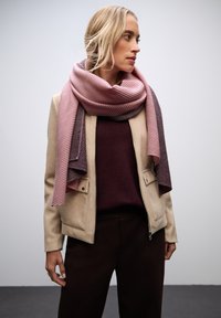 Giacca beige con chiusura a zip, maglione bordeaux sotto, e una sciarpa a righe rosa e grigie appesa al collo. Pantaloni marroni.