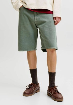 Person med olivengrønne knelang shorts, mørkebrune sokker og brune lær-laced sko, stående med den ene hånden i lommen.