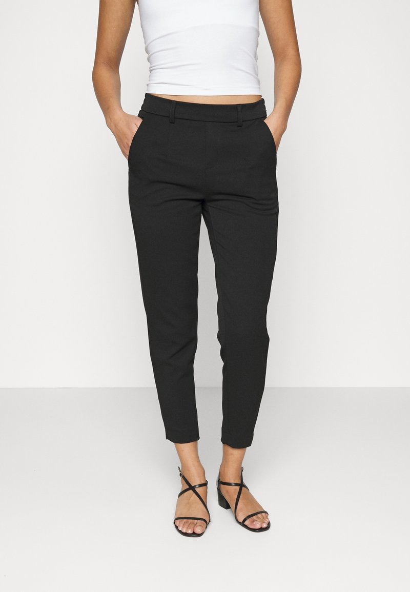 Pantalons noirs ajustés avec une coupe slim, présentant un devant plat et des passants de ceinture. Associés à des sandales à lanières noires et un débardeur blanc.