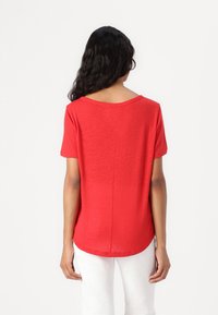 T-shirt rossa a maniche corte realizzata in tessuto leggero, con scollatura rotonda e orlo curvo sul retro, indossata con pantaloni bianchi.