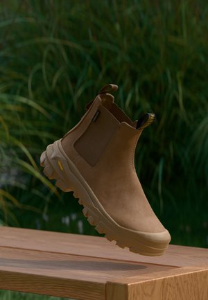 Bota de tobillo de cuero marrón con suela gruesa y estriada levitando sobre una superficie de madera con plantas verdes desenfocadas en el fondo.