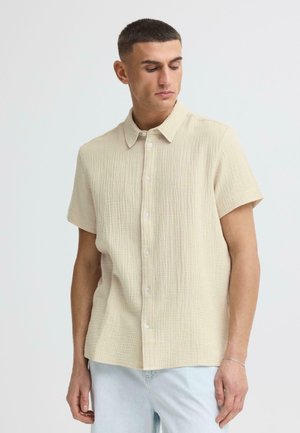 Mann mit kurzen dunklen Haaren, der ein beiges, strukturiertes Kurzarmhemd mit Knöpfen und hellblaue Jeans trägt und nach unten schaut.