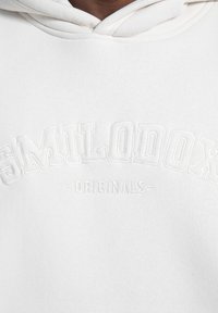 Sudadera blanca con el nombre de la marca "SMILODON" bordado en relieve en un diseño curvado, acompañado de "ORIGINALS" debajo, fabricada en suave material de algodón.
