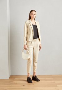 Blaser beige ajusté et joggers assortis en tissu doux, associés à un haut noir et des sabots foncés, tenant un sac à main crème. Fond en parquet.