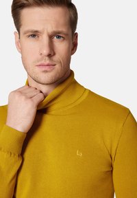 Musztardowy sweter typu golf w strukturze żebrowej z dopasowanym krojem. Posiada małe haftowane logo na piersi.