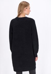 Vrouw met blond haar gezien van achteren, in een lange, zwarte, fuzzy trui-jurk en donkere broek, tegen een vlakke witte achtergrond.