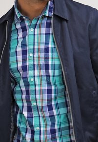 Man som bär en mörkblå jacka öppen över en teal, blå och vit rutig skjorta med knappar, där skjortans framsida och krage syns i detalj.