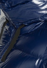 Glossy navy blue quilted jacket close-up showing padded texture, stitching details, and black drawstrings on the hood.