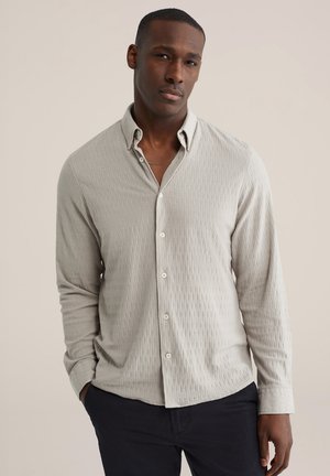 Lichtgrijs overhemd met lange mouwen en knoopsluiting, voorzien van een gestructureerd geribd patroon en knopen. Draagt donkere broek.
