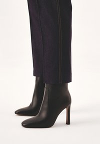 Black leather ankle boots with pointed toes and thin stiletto heels. Features seamless side panels and a smooth texture, paired with dark trousers.