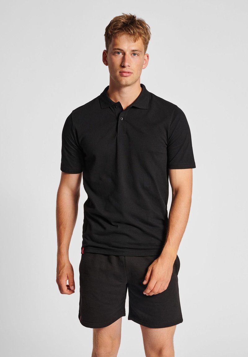 Junger Mann mit kurzen blonden Haaren, der ein eng anliegendes schwarzes Poloshirt und schwarze Shorts trägt und vor einem schlichten hellen Hintergrund steht.