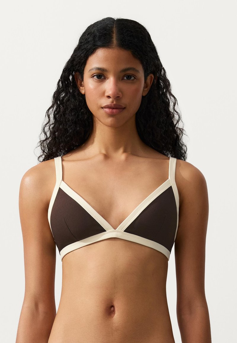 Femme aux cheveux bouclés foncés portant un soutien-gorge triangle marron avec une bordure crème, regardant vers l’avant sur un fond clair uni.