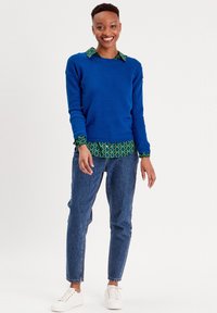 Pull en maille bleu sur chemise à motifs verts, associé à un jean bleu taille haute et des baskets blanches. Design simple et coupe décontractée.