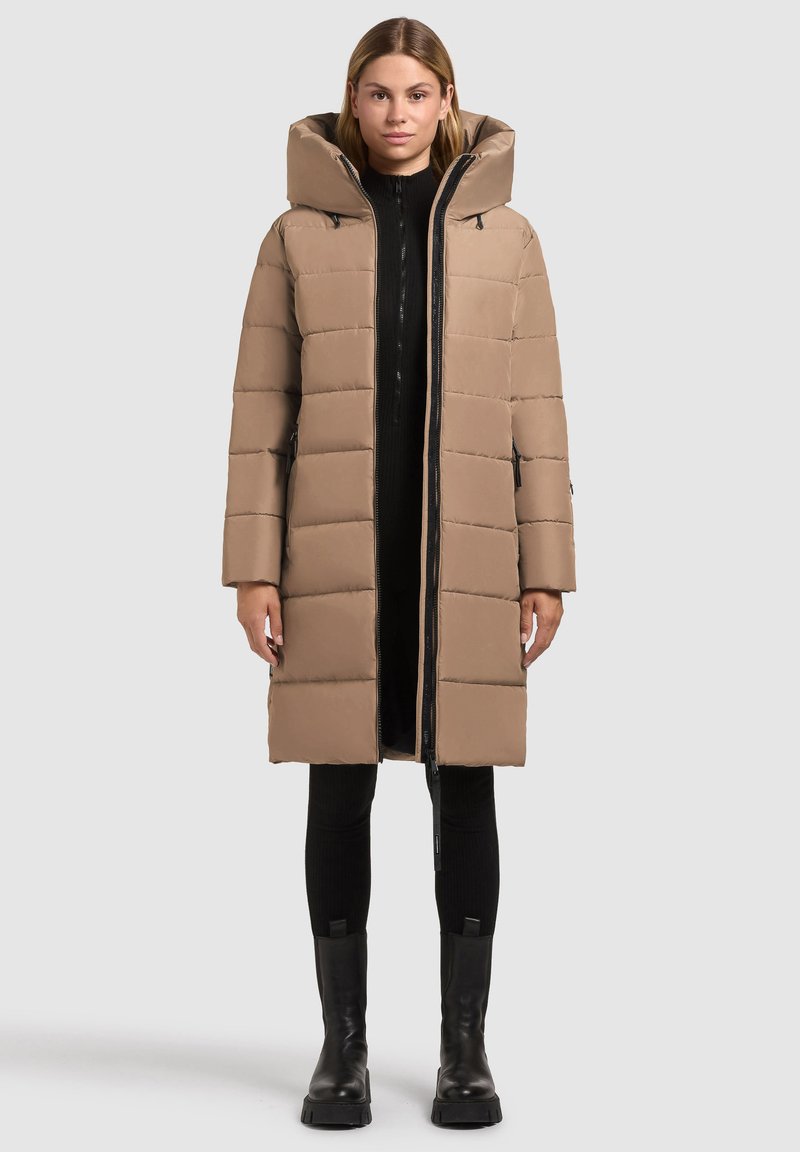 Lang, lys brun puffjakke med høj krave og lynlås, der har quilte sektioner og sidelommer. Båret over et sort outfit.