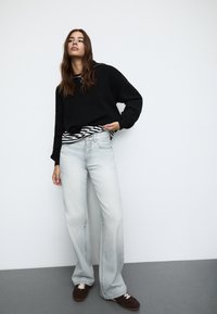 Črn pleten pulover preplaščan čez belo in črno črtasto srajco, svetlo prane široke hlače in rjave čevlje na vezanje na sivi podlagi.