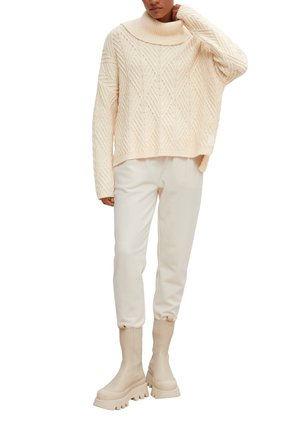 Personne portant un pull à col roulé en maille torsadée crème, un pantalon de jogging assorti et des bottines à plateforme épaisses, debout avec une main sur la nuque.