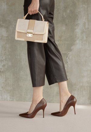 Scarpe con tacco alto in pelle marrone con punta affusolata, abbinate a pantaloni cropped in pelle scura e una borsa beige con un'accente testurizzato.