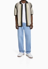 Camisa de punto de manga corta con rayas beige y verdes, collar negro, llevada sobre una camiseta blanca, combinada con unos jeans azul claro y zapatillas blancas.